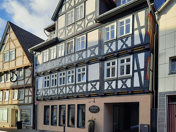 Fachwerkhaus im Landkreis Hersfeld-Rotenburg. Das mehrstöckige Gebäude besitzt eine aufwendig verzierte Fassade mit dunklen Holzstreben und weißem Putz. Die untere Etage wurde modern renoviert, während die oberen Stockwerke traditionelle Fachwerk-Elemente aufweisen. Im Hintergrund sind weitere Fachwerkhäuser und ein blauer Himmel mit einigen Wolken zu sehen.